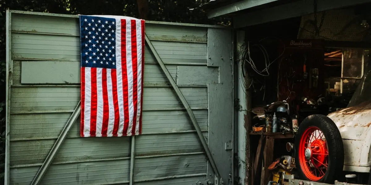 American flag hanging on a weathered storage shed beside an old garage with a vintage car wheel.