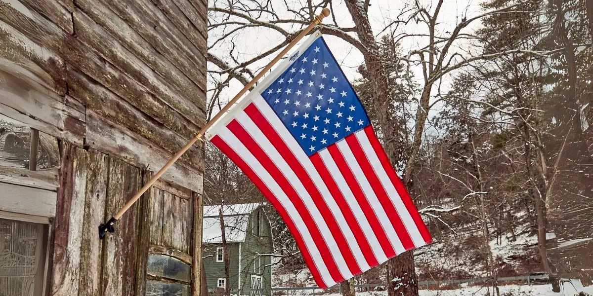 American flag mounted on a rustic wooden building, waving in a snowy winter landscape with trees and houses in the background.