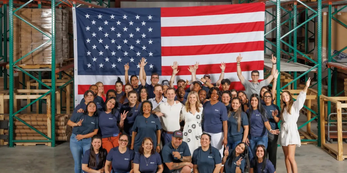 American flag store team posing in front of a large American flag inside a warehouse with shelves and packaging materials.