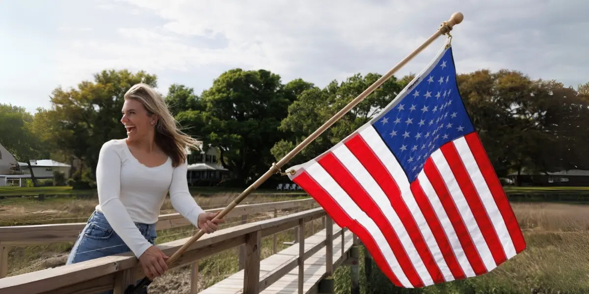 High-quality U.S. flag waving as a smiling woman holds it on a wooden dock with greenery and houses in the background.