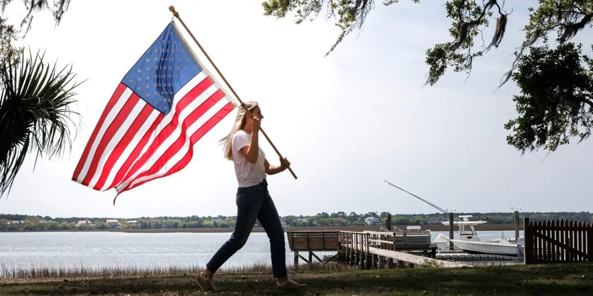 High quality American flag waving as a woman walks by a waterfront dock with boats in the background.